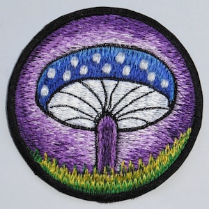 Könnte beinhalten: Gestickter Aufnäher mit einem lila und blauen Pilz mit weißen Punkten auf dem Hut. Der Pilz ist von grünem Gras umgeben.
