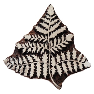 May include: A wooden printing block featuring a fern leaf design. The leaf pattern is white against a dark brown background. The block is triangular, with the fern design carved into the wood, creating a textured surface.
