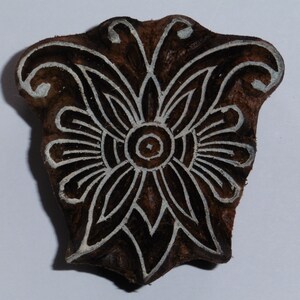 Könnte beinhalten: Ein Holzblock-Stempel mit einem detaillierten floralen Design. Das Design zeigt eine zentrale Blume mit filigranen Blütenblättern und einem kleineren Kreis in der Mitte. Der Stempel ist braun mit weißen Linien, die das Design umranden.