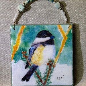 May include: Square ceramic ornament featuring a detailed watercolor painting of a chickadee perched on a pine branch. The bird has a black head, white cheeks, and yellow breast. The ornament has a beaded loop for hanging and the initials "K.J.T." are visible.