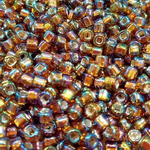 May include: A close-up image of a large quantity of iridescent glass seed beads. The beads are a mix of orange, brown, and gold colours with a metallic sheen.