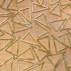 May include: A collection of gold-coloured triangle charms with small loops for jewellery making. The charms are scattered on a textured orange background. The triangles are all the same size and shape.