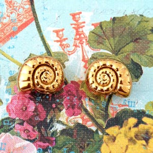 May include: Two gold-coloured seashell stud earrings. Each earring is shaped like a spiral shell with detailed ridges. The background has a floral design with pink, yellow, and green elements.