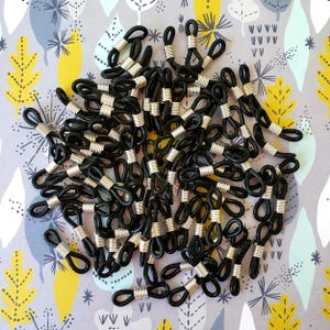 May include: A pile of black spectacle retainers with silver metal coils. The retainers are made of a flexible material and are designed to loop around the arms of spectacles to keep them secure. The background is a patterned fabric.