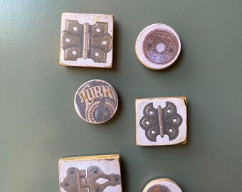 Könnte beinhalten: Sechs dekorative Magnete mit verschiedenen Designs, darunter Scharniere, ein Zifferblatt und florale Muster. Die Magnete sind alle quadratisch oder rund und haben einen weißen Hintergrund mit braunen und goldenen Akzenten.