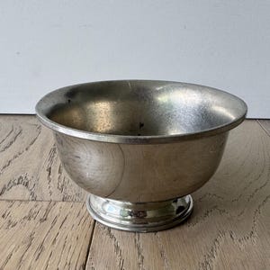 May include: A silver-coloured metal bowl with a rounded body and a pedestal base. The bowl has a wide opening and a smooth, reflective surface. The bowl is likely used for serving or display.