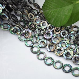 May include: A collection of iridescent, ring-shaped beads with a mix of colors including green, blue, and brown. The beads are arranged in a pile, with some in focus and others blurred. A green leaf is visible in the background.