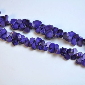 May include: Two strands of purple, irregularly shaped beads. The beads have a polished, glossy finish and are strung together, creating a necklace or bracelet. The background is a plain white surface.