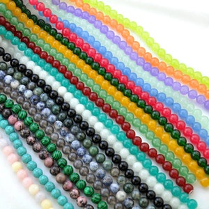 Puede incluir: Surtido de cuentas redondas de piedras preciosas en varios colores, incluyendo turquesa, verde, rojo, amarillo, azul y blanco. Las cuentas est&aacute;n enhebradas sobre un fondo blanco.