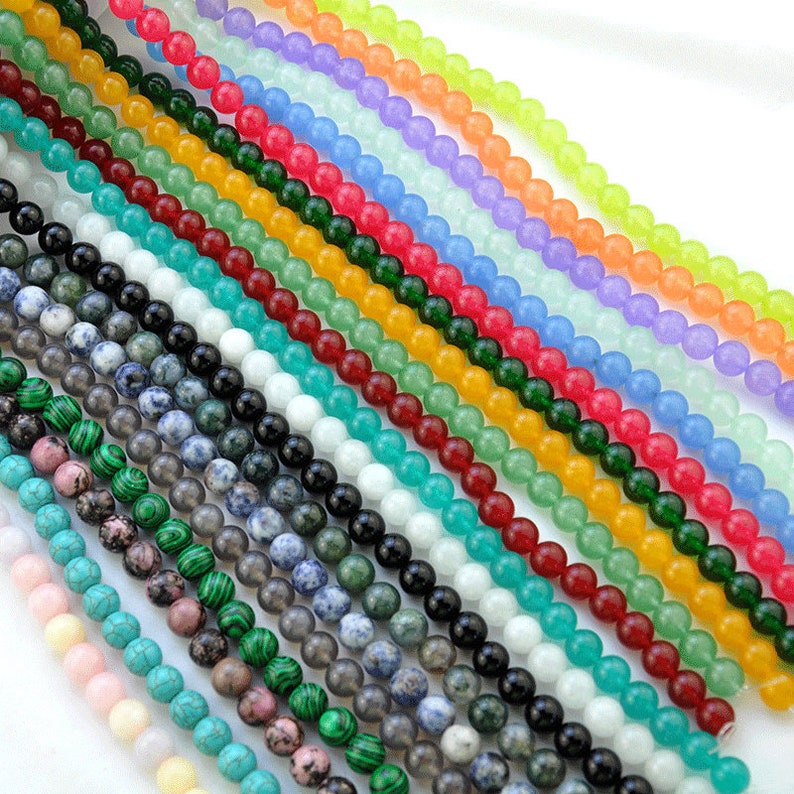 Puede incluir: Surtido de cuentas redondas de piedras preciosas en varios colores, incluyendo turquesa, verde, rojo, amarillo, azul y blanco. Las cuentas est&aacute;n enhebradas sobre un fondo blanco.