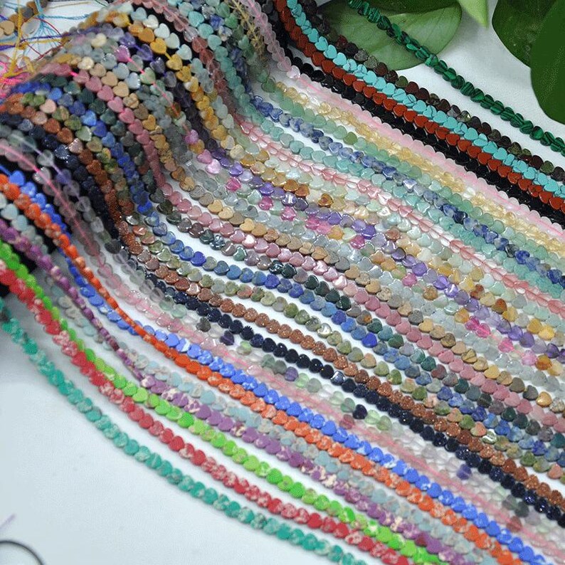 Puede incluir: Una colecci&oacute;n de cuentas en forma de coraz&oacute;n en varios colores, incluyendo rosa, azul, verde y marr&oacute;n. Las cuentas est&aacute;n ensartadas, creando m&uacute;ltiples hebras. La imagen muestra una variedad de cuentas de piedras preciosas, perfectas para la creaci&oacute;n de joyas.