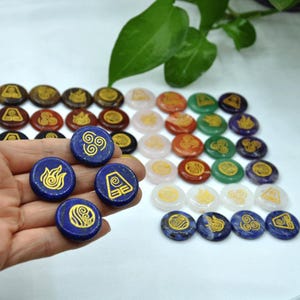 May include: Set of 25 gemstone coasters with gold painted symbols. The symbols are the four elements from Avatar: The Last Airbender. The coasters are in various colours, including pink, green, red, purple, and blue.