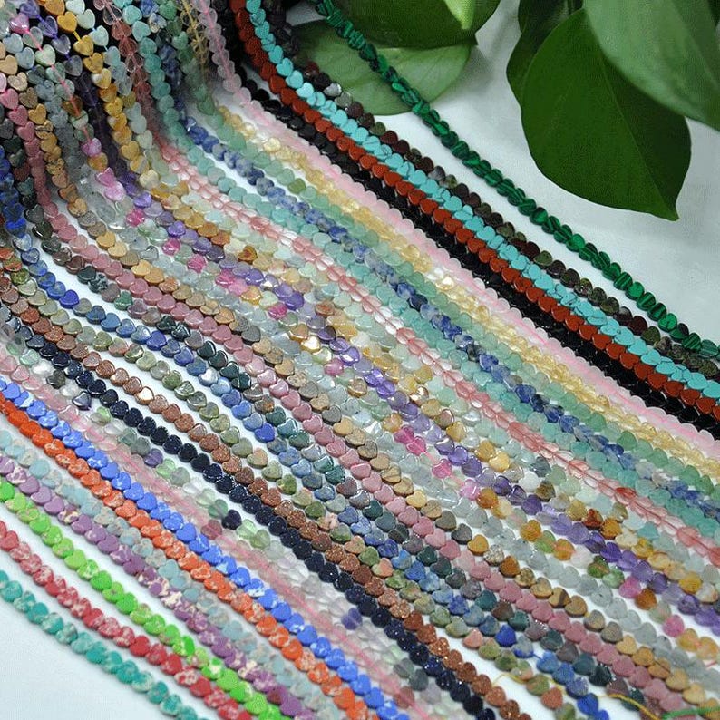 Puede incluir: Una colecci&oacute;n de cuentas de piedras preciosas en forma de coraz&oacute;n en varios colores, incluyendo verde, azul, rosa y marr&oacute;n. Las cuentas est&aacute;n ensartadas, creando una exhibici&oacute;n texturizada y colorida. Las cuentas son probablemente para hacer joyas.