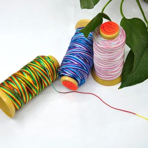 May include: Three spools of colorful thread. One spool is multi-colored with yellow, green, red, and orange. Another is blue and purple. The third is pink and white. All spools are on a yellow base.