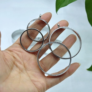 Könnte beinhalten: Vier transparente Glas- und silberfarbene Metall-Kreisanhänger mit kleinen Schlaufen oben. Die Anhänger werden in einer Hand gehalten, mit einem unscharfen silberfarbenen Metallkreis im Hintergrund. Die Anhänger sind wahrscheinlich für die Schmuckherstellung oder das Basteln.