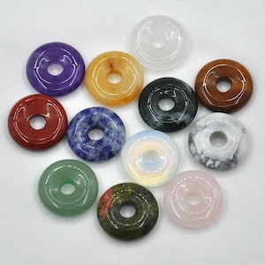 Pode incluir: Doze contas de pedras preciosas em forma de rosquinha em v&aacute;rias cores, incluindo ametista, laranja, preto, branco, azul, verde, vermelho e rosa. As contas est&atilde;o dispostas em um c&iacute;rculo sobre uma superf&iacute;cie branca.