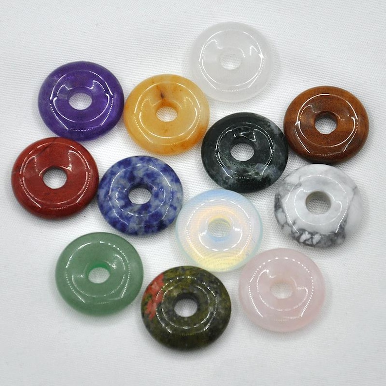 Pode incluir: Doze contas de pedras preciosas em forma de rosquinha em v&aacute;rias cores, incluindo ametista, laranja, preto, branco, azul, verde, vermelho e rosa. As contas est&atilde;o dispostas em um c&iacute;rculo sobre uma superf&iacute;cie branca.