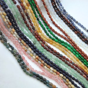 Pu&ograve; includere: Assortimento di perline di pietre preziose di forma ovale in vari colori, tra cui verde, blu, marrone, rosa e giallo. Le perline sono infilate su uno sfondo bianco.