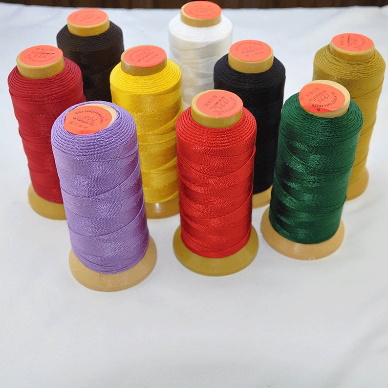 May include: A collection of colorful spools of thread in various shades, including red, purple, yellow, and green. Each spool is wound tightly and has a wooden base and a paper label on top. The threads appear to be made of a shiny, smooth material.