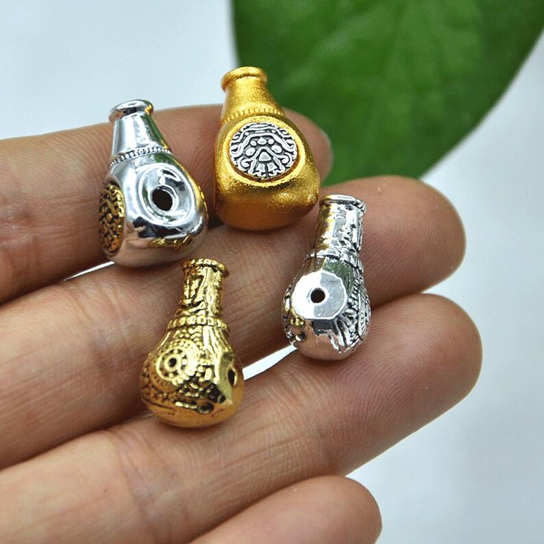 May include: Four small, decorative bottle-shaped beads in gold and silver tones. The beads have intricate patterns and a hole for stringing. The gold beads have a shiny finish, while the silver beads have a matte finish.