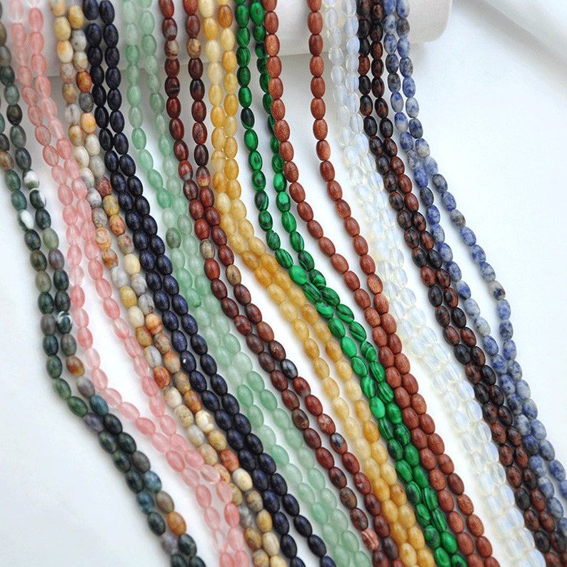 Pu&ograve; includere: Una variet&agrave; di perline di pietre preziose di forma ovale in diversi colori, tra cui verde, blu, marrone, rosa e giallo. Le perline sono infilate su uno sfondo bianco.