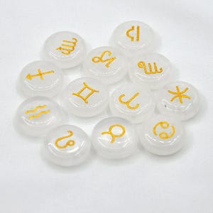 May include: Twelve white, round, smooth stones with gold zodiac symbols. The symbols are: Aries, Taurus, Gemini, Cancer, Leo, Virgo, Libra, Scorpio, Sagittarius, Capricorn, Aquarius, and Pisces.