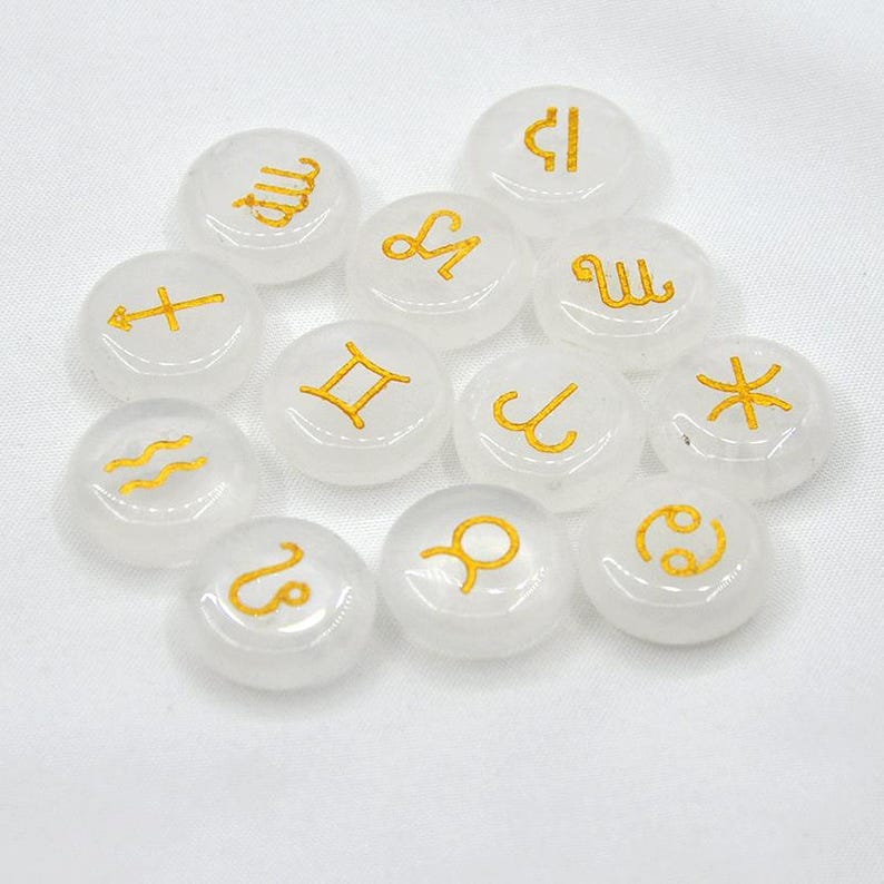 May include: Twelve white, round, smooth stones with gold zodiac symbols. The symbols are: Aries, Taurus, Gemini, Cancer, Leo, Virgo, Libra, Scorpio, Sagittarius, Capricorn, Aquarius, and Pisces.