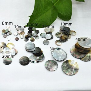 Op de afbeelding: Verschillende maten ronde parelmoer schelpkralen, variërend van 8 mm tot 40 mm in diameter. De kralen hebben tinten van wit, grijs en bruin, met een natuurlijke, parelmoerglans. Een groen blad is zichtbaar op de achtergrond.