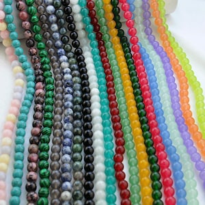 Puede incluir: Surtido de cuentas redondas de piedras preciosas en varios colores, incluyendo rosa, verde, azul, turquesa y amarillo. Las cuentas est&aacute;n enhebradas sobre un fondo blanco.