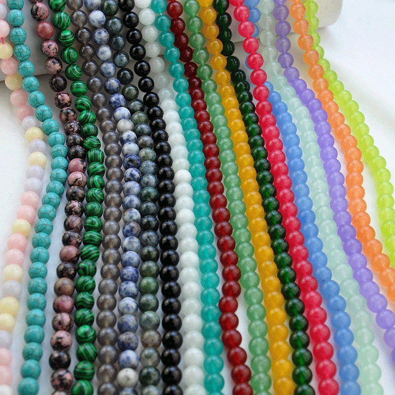 Puede incluir: Surtido de cuentas redondas de piedras preciosas en varios colores, incluyendo rosa, verde, azul, turquesa y amarillo. Las cuentas est&aacute;n enhebradas sobre un fondo blanco.