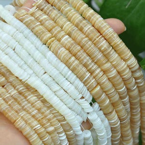 May include: Multiple strands of flat, circular disc beads arranged in parallel rows. The beads feature a smooth, polished finish with a pearlescent sheen, alternating between solid white and light tan or beige color variations throughout the collection.