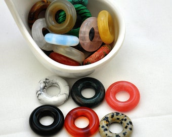 May include: Assortment of colorful gemstone rings in a white ceramic bowl. The rings are made of various stones, including turquoise, agate, and jasper.