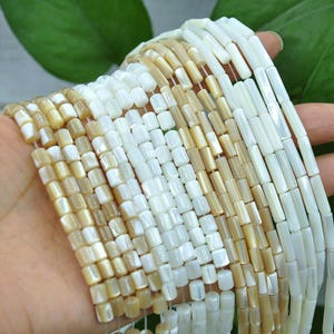 May include: Multiple strands of cylindrical beads made from iridescent mother-of-pearl shell. The beads feature a smooth, polished finish with a natural colour gradient ranging from creamy beige and light tan to bright, reflective white tones.