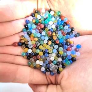 May include: A collection of colorful gemstone beads in various shades of blue, green, red, yellow, and brown. The beads are small and round, and they are arranged in a pile in the palm of a hand.