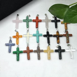 May include: A selection of gemstone cross pendants in diverse colours, such as pink, green, orange, white, black, and brown. Each cross features a silver-coloured bail for attaching to a chain. The crosses are presented against a white backdrop.