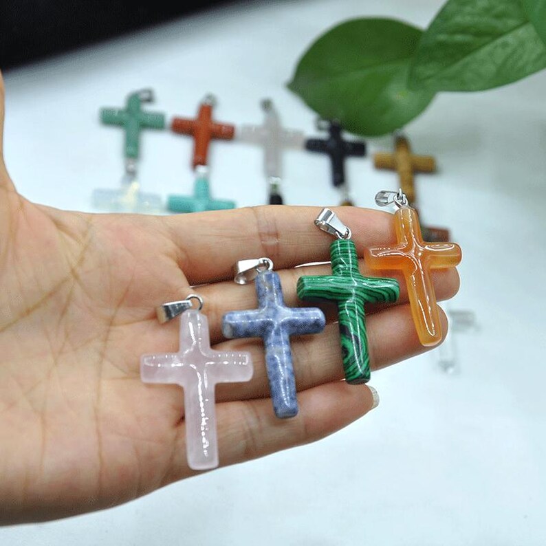May include: A selection of gemstone cross pendants in shades of pink, blue, green, and orange. Each cross features a silver-coloured bail for attaching to a chain. The pendants are crafted from polished stone and are presented against a soft backdrop.