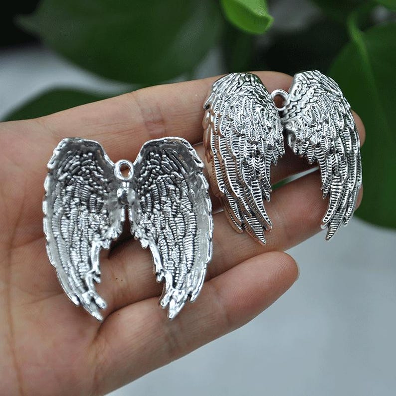 May include: Two silver-colored angel wing charms. The charms are detailed with feather-like textures and have a loop at the top for attaching to jewelry or other crafts. The wings are displayed against a blurred background.