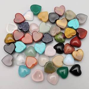 Peut inclure: Assortiment de pierres polies en forme de cœur de différentes couleurs, notamment rouge, vert, bleu, noir, blanc et marron. Les pierres sont disposées de manière aléatoire sur un fond blanc.