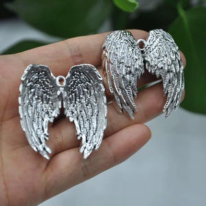 May include: Silver-colored metal angel wing charms. The charms have detailed feather textures and a loop at the top for attaching to jewelry or crafts. The wings are displayed against a neutral background.
