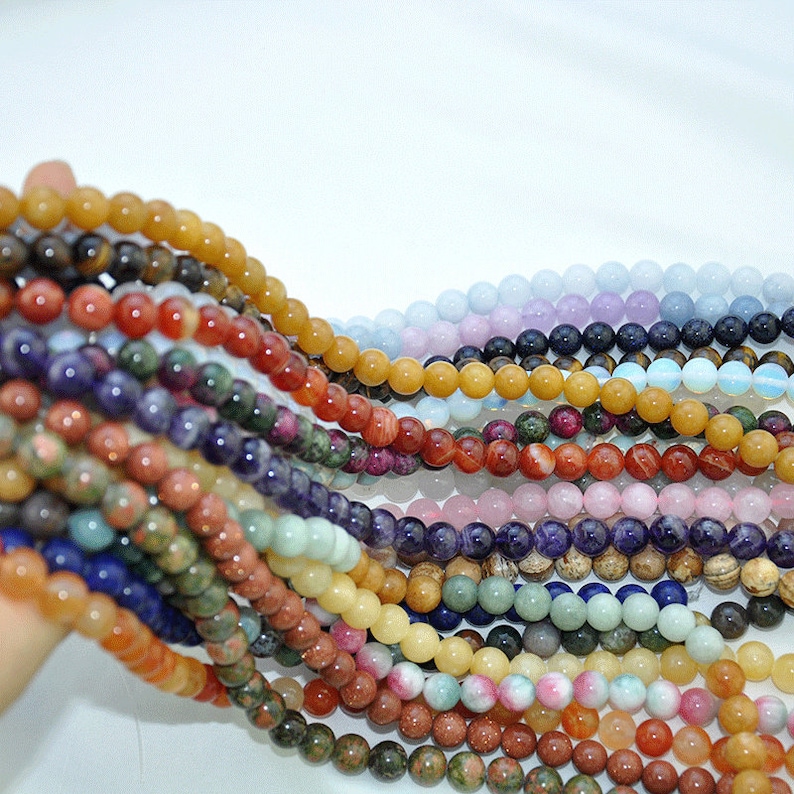 Puede incluir: Una variedad de cuentas redondas de piedras preciosas en diferentes colores, incluyendo amarillo, naranja, rojo, verde, azul, morado y blanco. Las cuentas est&aacute;n enhebradas en una larga hebra.