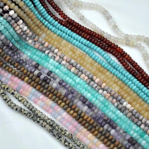 Puede incluir: Surtido de cuentas de piedras preciosas en varios colores y patrones, incluyendo turquesa, marrón, rosa, blanco y verde. Las cuentas están enhebradas sobre un fondo blanco.