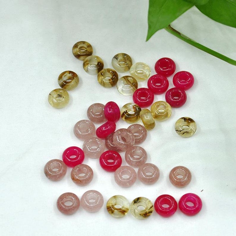May include: Assortment of donut-shaped beads in various shades of pink, brown, and clear. The beads are made of glass or stone and have a smooth, polished finish.