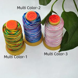 May include: Three spools of multi-colored thread. The spools are labeled "Multi Color-1", "Multi Color-2", and "Multi Color-3". The thread colors include rainbow, blue, purple, white, and pink. Each spool has a yellow base and an orange top.