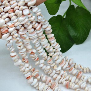 Peut inclure: Plusieurs brins de petites perles de coquillages blanc et pêche, de forme irrégulière et polies. Chaque perle présente une surface lisse avec des variations naturelles de forme et de couleur, enfilées pour former de longues lignes organiques.