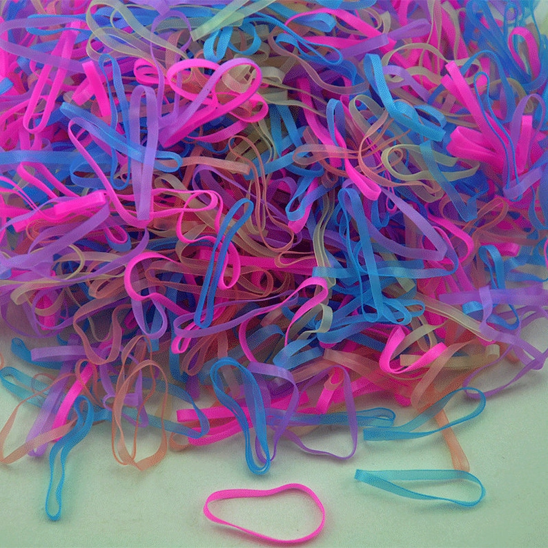 Pu&ograve; includere: Un grande mucchio di elastici colorati in rosa, blu e trasparente. Gli elastici sono disposti in modo casuale e sono tutti della stessa dimensione.