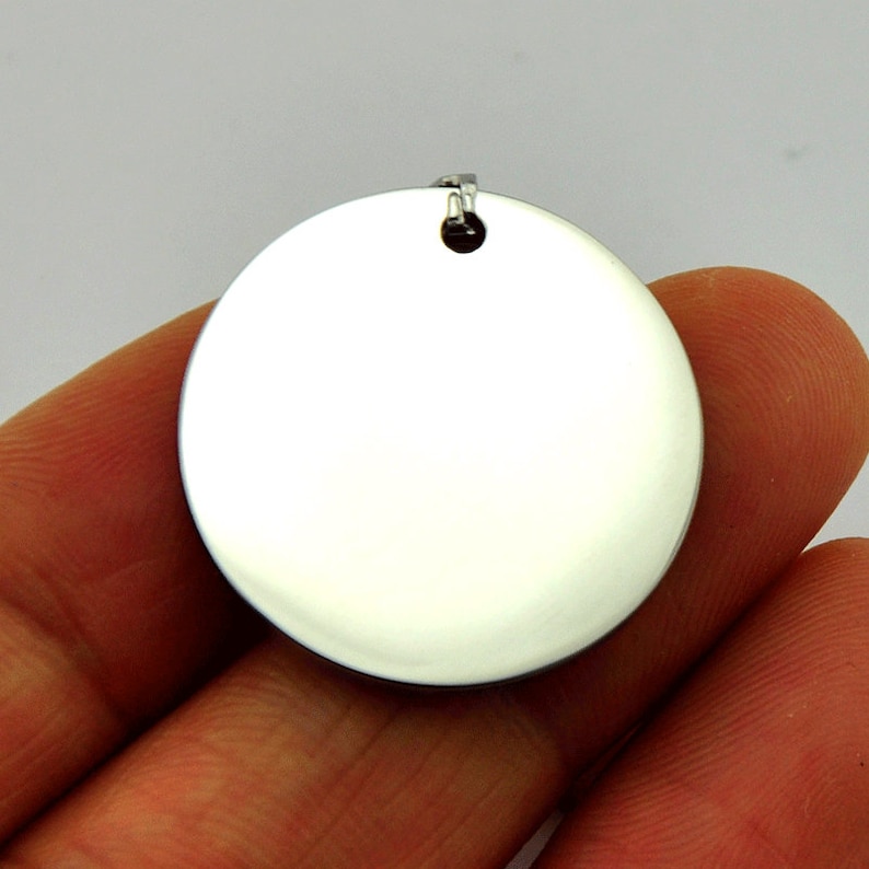 May include: A round, silver-colored pendant with a smooth, shiny surface. The pendant has a small loop at the top for attaching a chain.