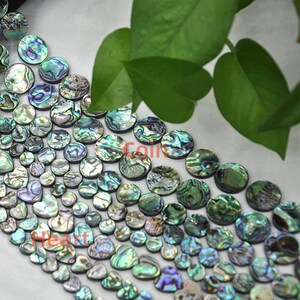 Könnte beinhalten: Eine Sammlung schillernder Abalone-Muschelkernperlen in verschiedenen Formen und Größen. Die Perlen zeigen eine Reihe von Farben, darunter Grün, Blau und Lila, mit einem natürlichen, strukturierten Aussehen. Einige Perlen sind rund, andere herzförmig.