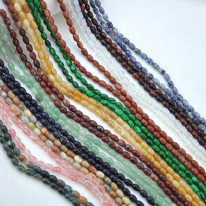 Pu&ograve; includere: Assortimento di perline di pietre preziose di forma ovale in vari colori, tra cui verde, blu, marrone, giallo e rosa. Le perline sono infilate su uno sfondo bianco.