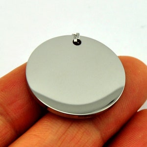 May include: A round, silver-colored pendant with a smooth, polished surface. The pendant has a small loop at the top for attaching a chain.