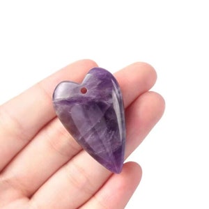 Puede incluir: Un colgante de corazón de amatista púrpura con un agujero para un cordón o cadena. El colgante es liso y pulido.